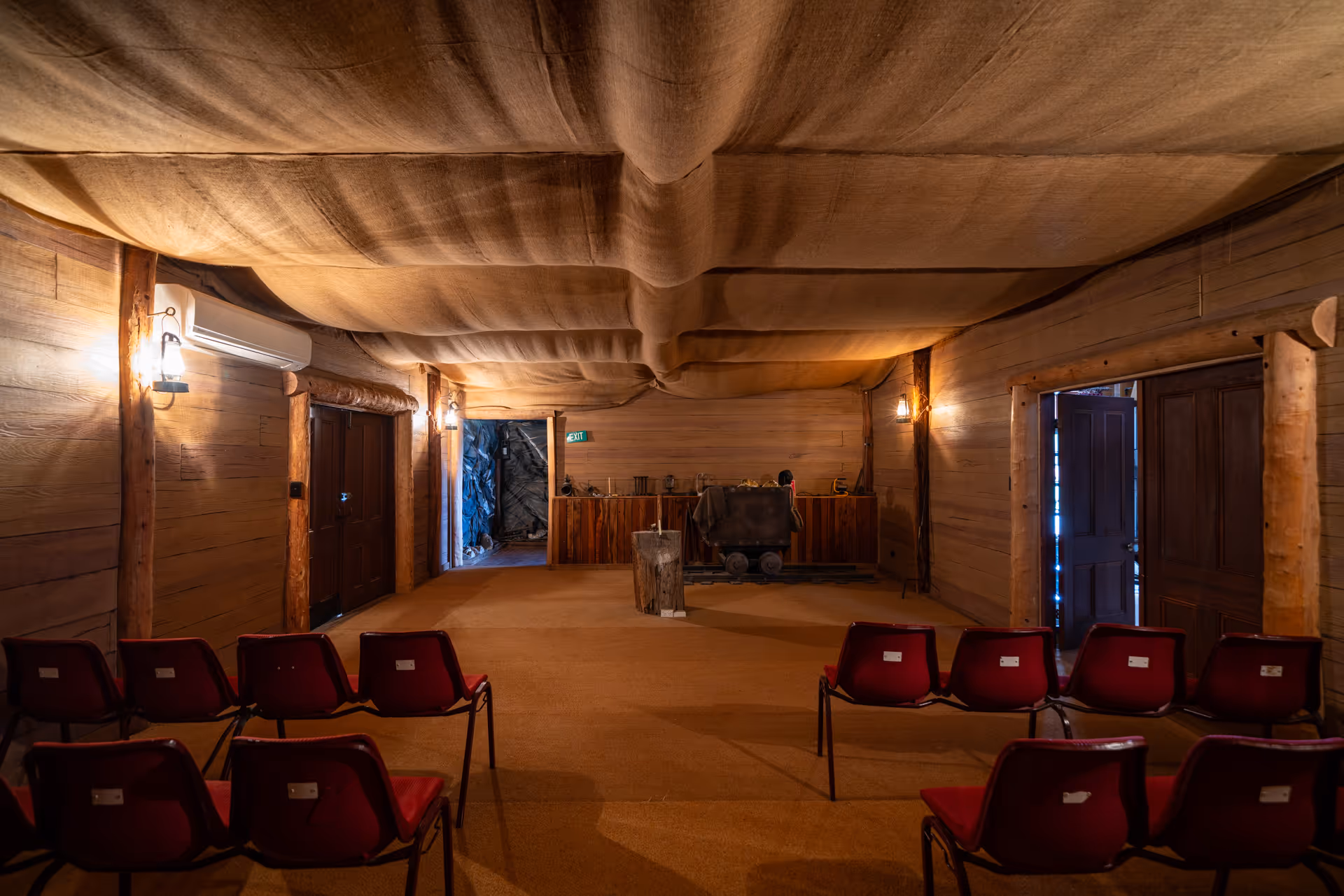 Interior of a wooden room with draped ceiling, rows of red chairs, wall lamps, and a small mining cart on tracks near an exit door.
