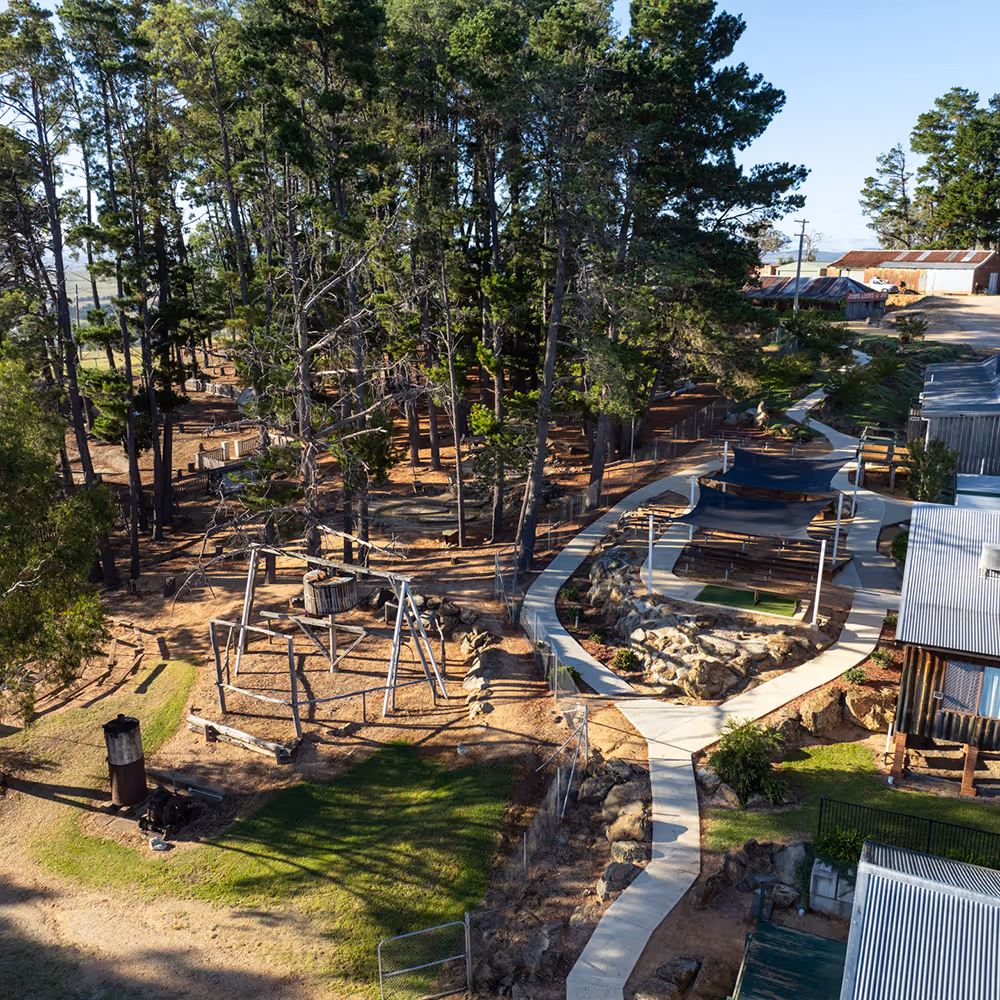 Aerial view of a forested camp area with wooden playground equipment, winding paved pathways, and shaded picnic tables.