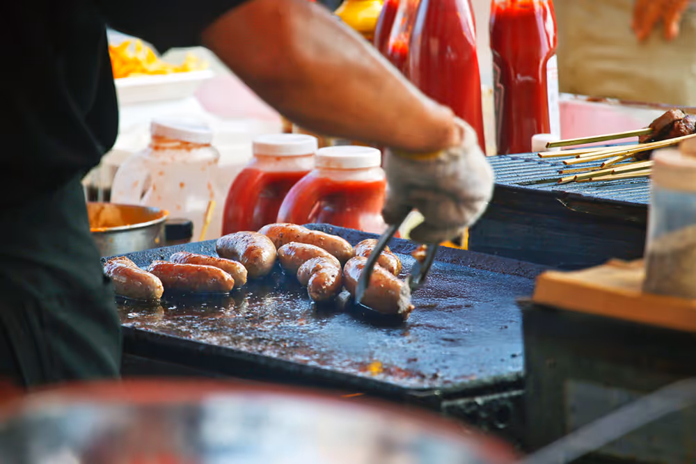 Person grilling sausages on a flat top grill with bottles of condiments in the background.