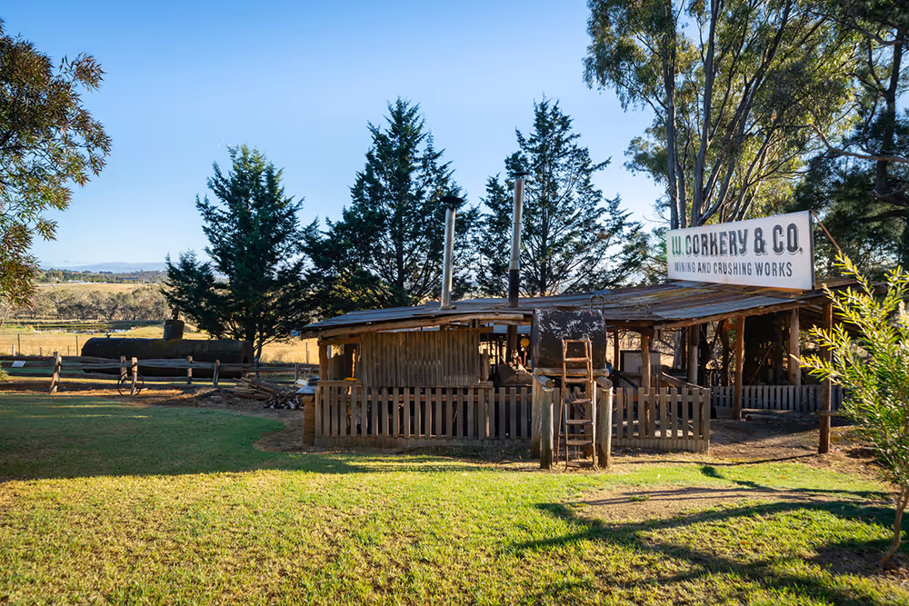Rustic wooden mining and crushing works building with a sign reading 'W. Corkery & Co. Mining and Crushing Works' surrounded by trees and grassy field under a clear blue sky.