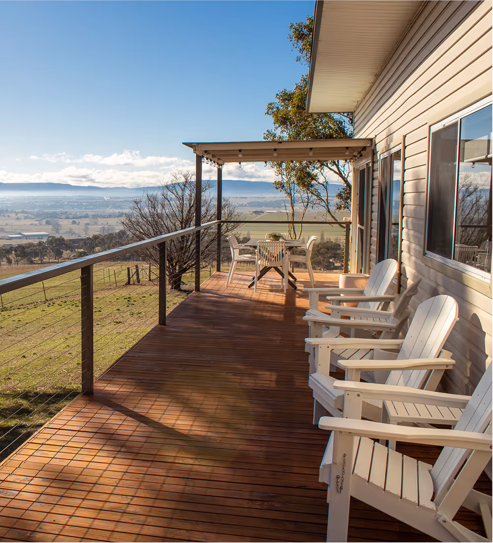 Wooden deck with white Adirondack chairs and a table under a pergola overlooking a scenic countryside landscape with hills and trees.