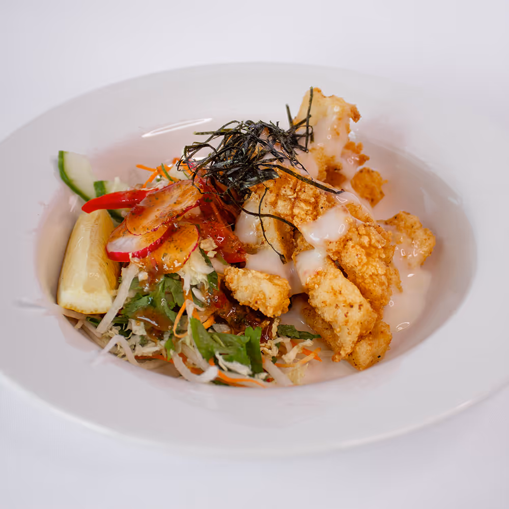 Plate with crispy fried fish topped with a creamy sauce, served with fresh mixed salad and a lemon wedge.