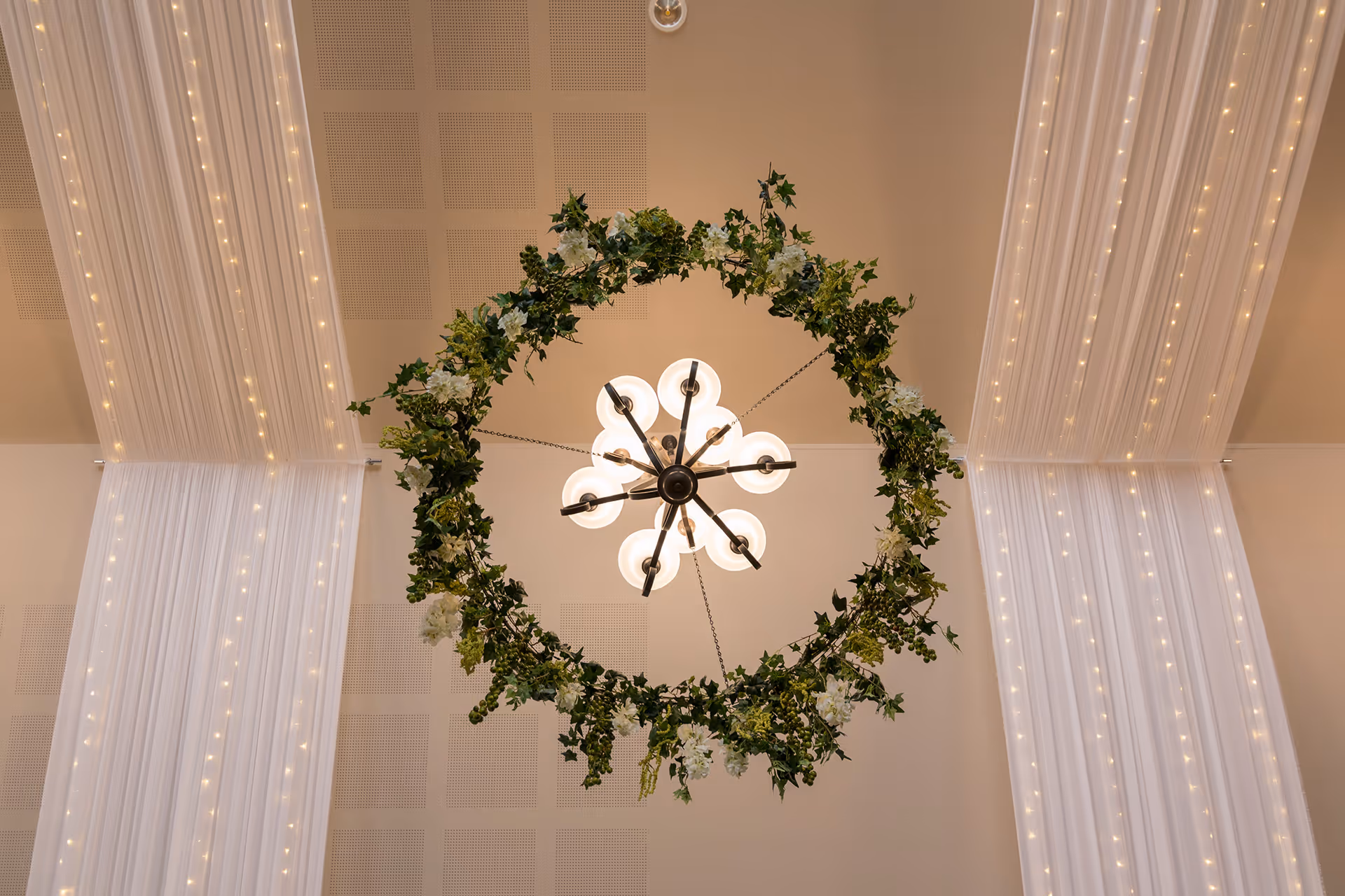 Ceiling chandelier with eight round lights surrounded by a circular wreath of green leaves and white flowers, with sheer white drapes and string lights in the background.