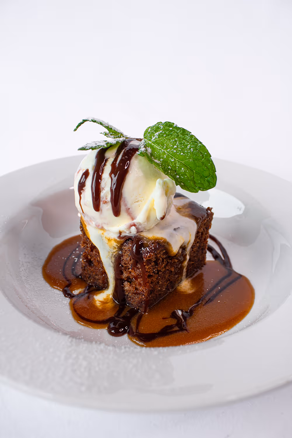 Chocolate cake topped with melting vanilla ice cream, drizzled chocolate sauce, and garnished with a mint leaf on a white plate.