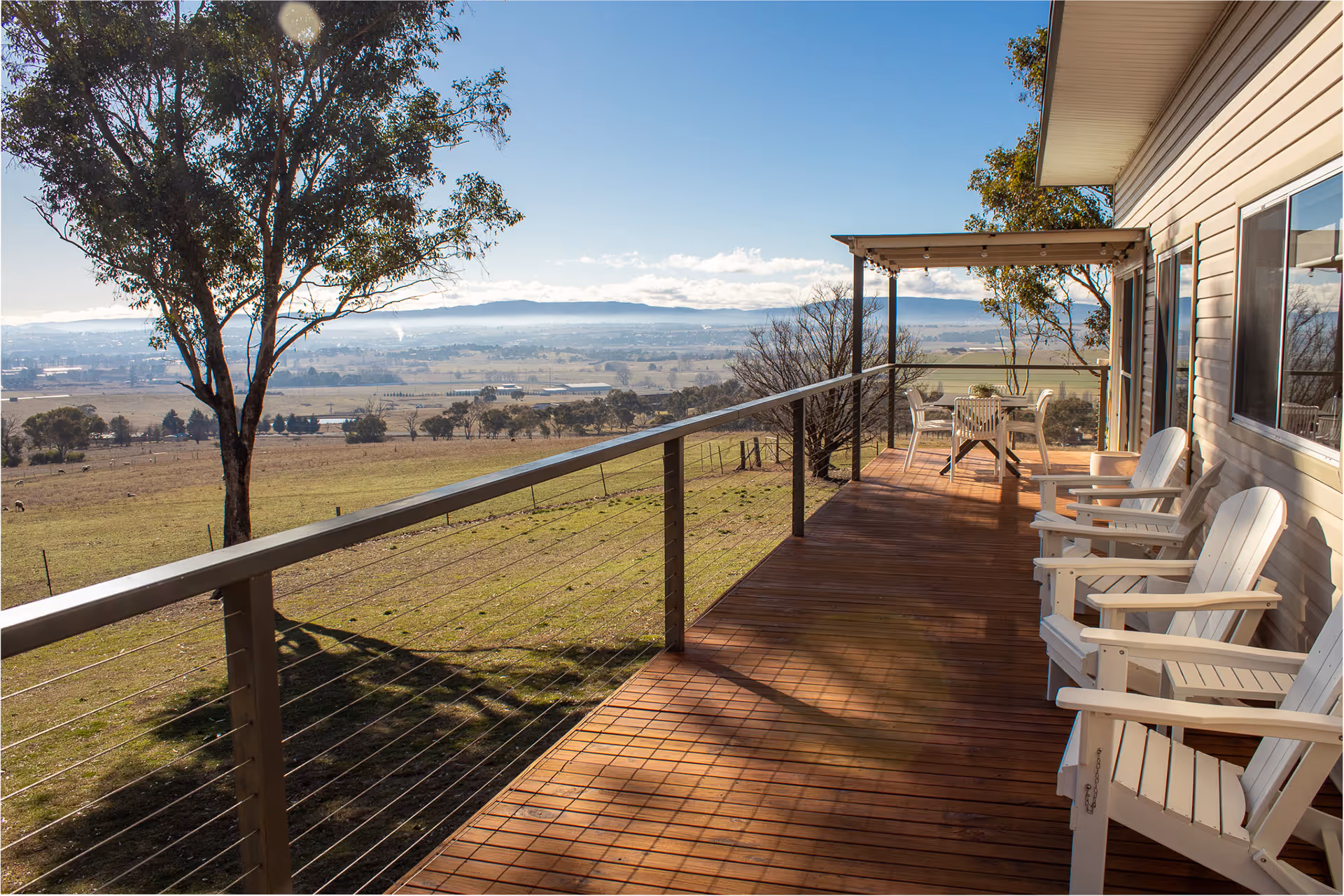 Wooden porch with white Adirondack chairs and table under a pergola, overlooking a scenic rural landscape with trees and distant hills.