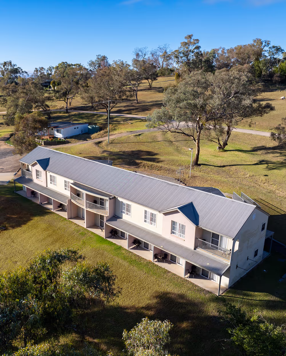 A long, two-story building with a gray corrugated roof, surrounded by grassy fields and scattered trees under a clear blue sky.
