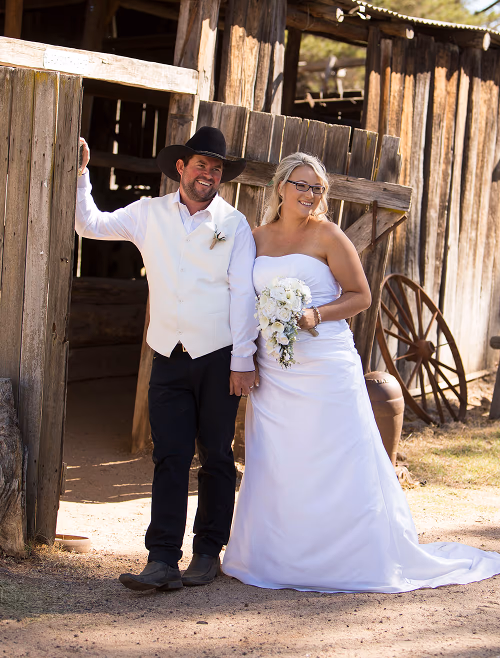 Smiling bride in white dress holding bouquet and groom in white vest and black cowboy hat standing by rustic wooden barn door.
