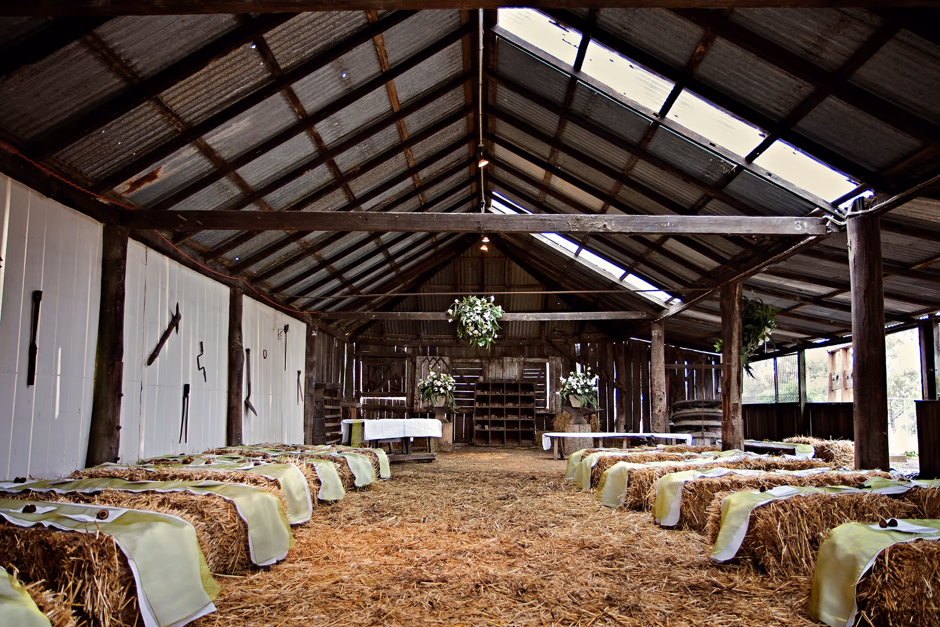 Rustic barn interior set up for a wedding with hay bale seating covered by pale green cloths and floral arrangements at the front.