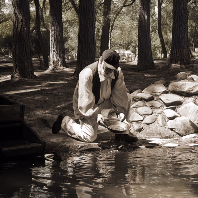 Man kneeling by a riverbank panning for gold with a pan during the gold rush era, surrounded by trees and rocks.