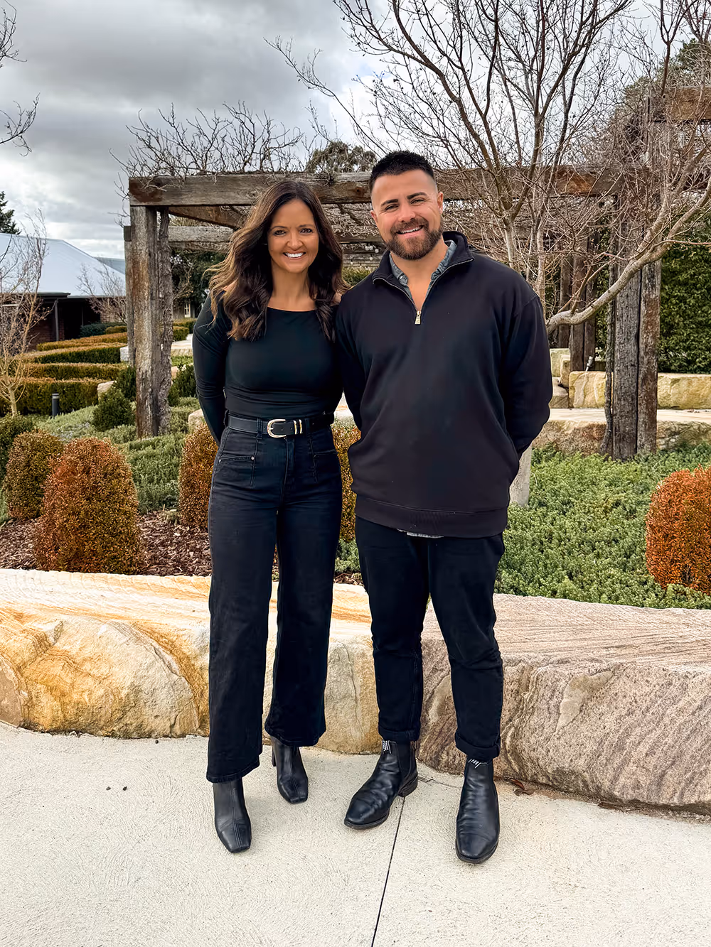 Smiling man and woman standing together outdoors in front of a stone bench and landscaped garden.