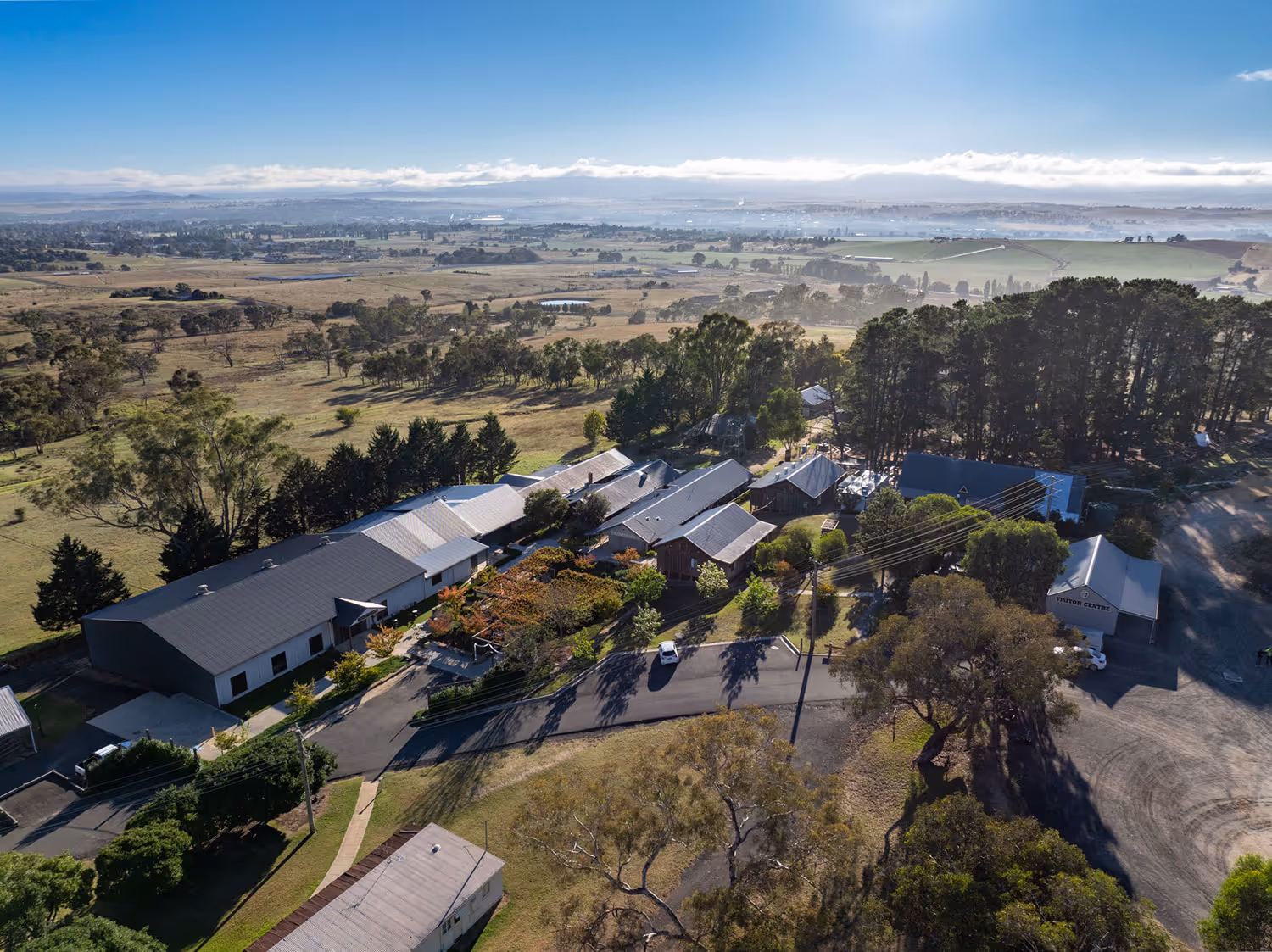 Aerial view of a rural property with several buildings surrounded by trees and open fields under a clear blue sky.