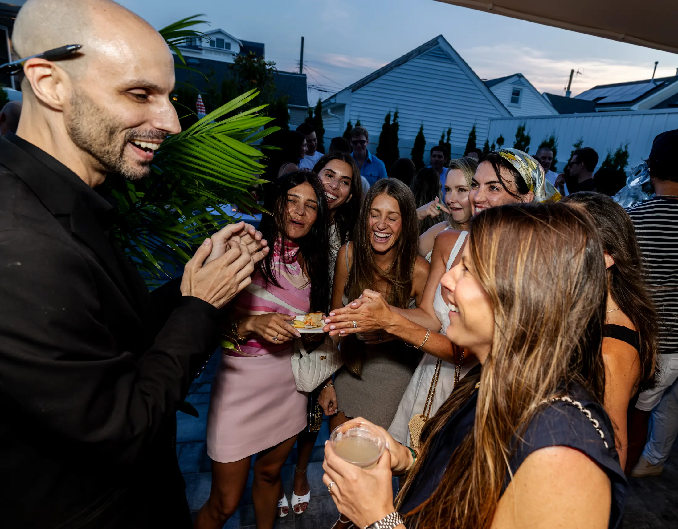 Magician Jesse Dameron in all-black business attire performing close-up magic for a group of laughing guests at a private summer party
