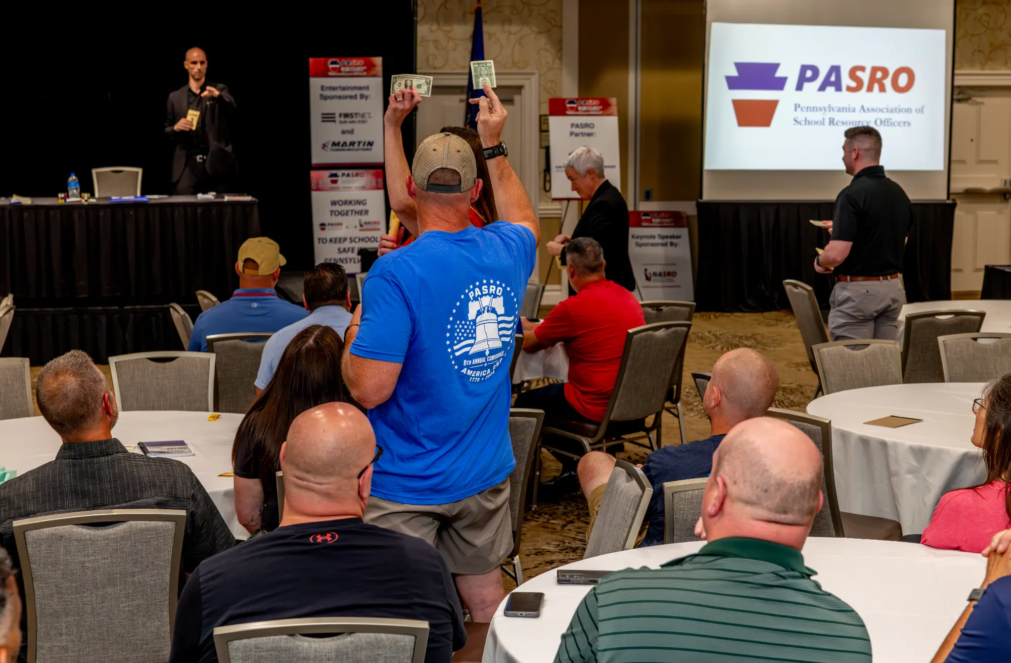Conference attendee standing among banquet tables holding two dollar bills in the air
