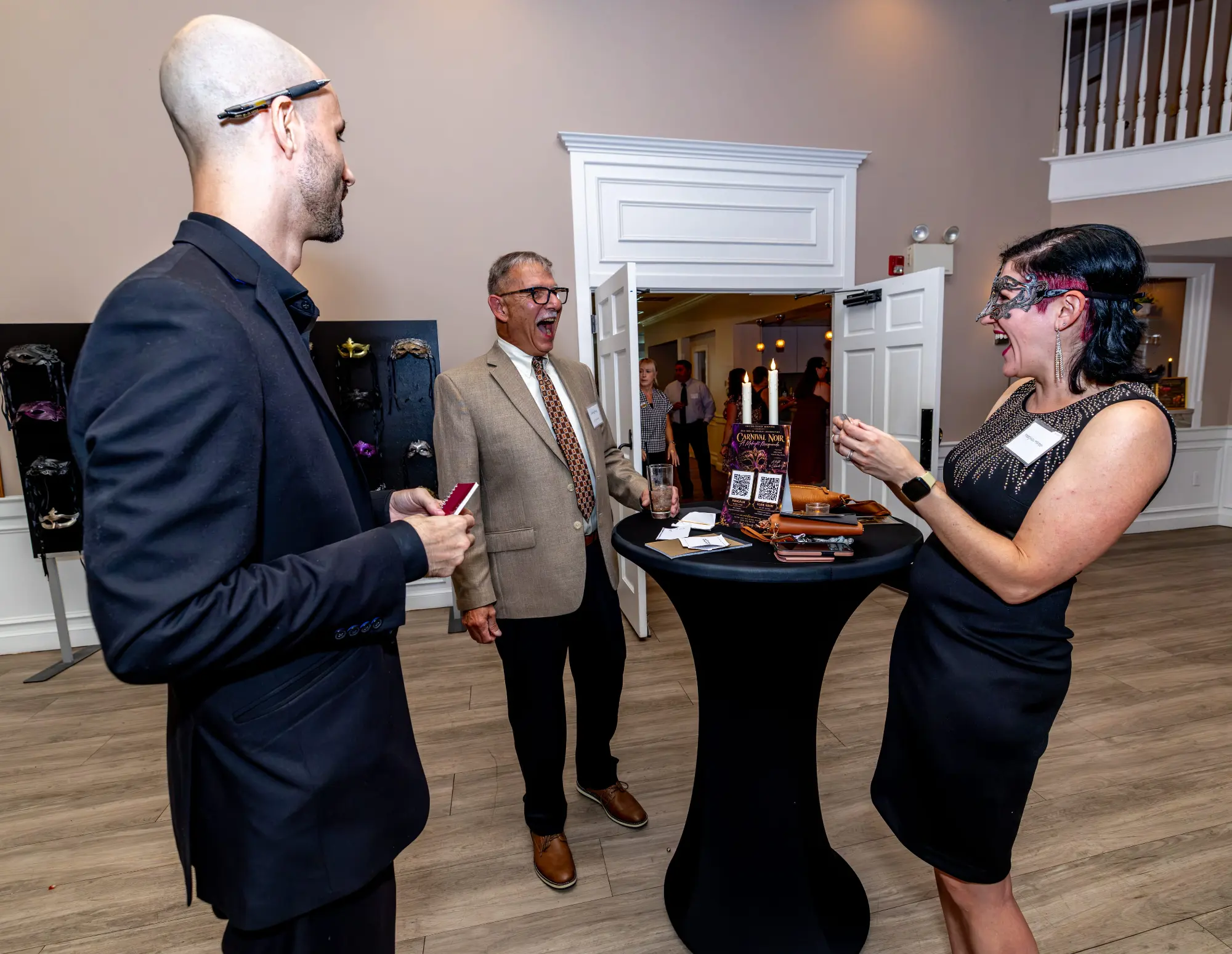 Jesse Dameron, magician and mentalist, with two laughing guests at a cocktail table beside a masquerade mask display