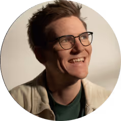 Smiling young man with short hair, wearing glasses and a light jacket, looking to the side.