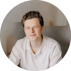 Young man with short curly hair wearing a light-colored shirt sitting on a chair.