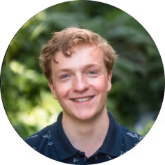 Smiling young man with light brown hair wearing a dark blue shirt against a blurred green outdoor background.