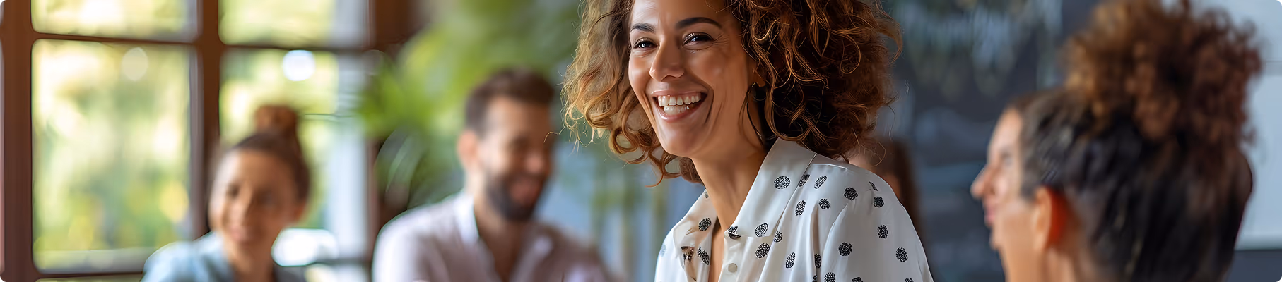 Mujer sonriente con cabello rizado con camisa blanca con patrones negros, sentada con gente borrosa en el fondo.