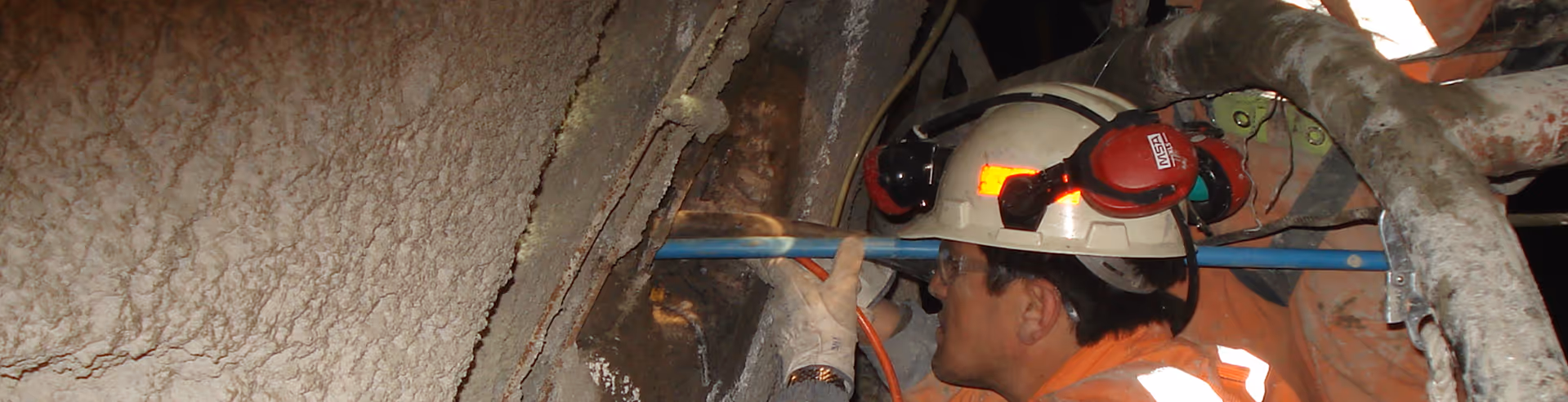 Trabajador con casco de seguridad y gafas operando maquinaria en un espacio industrial confinado.