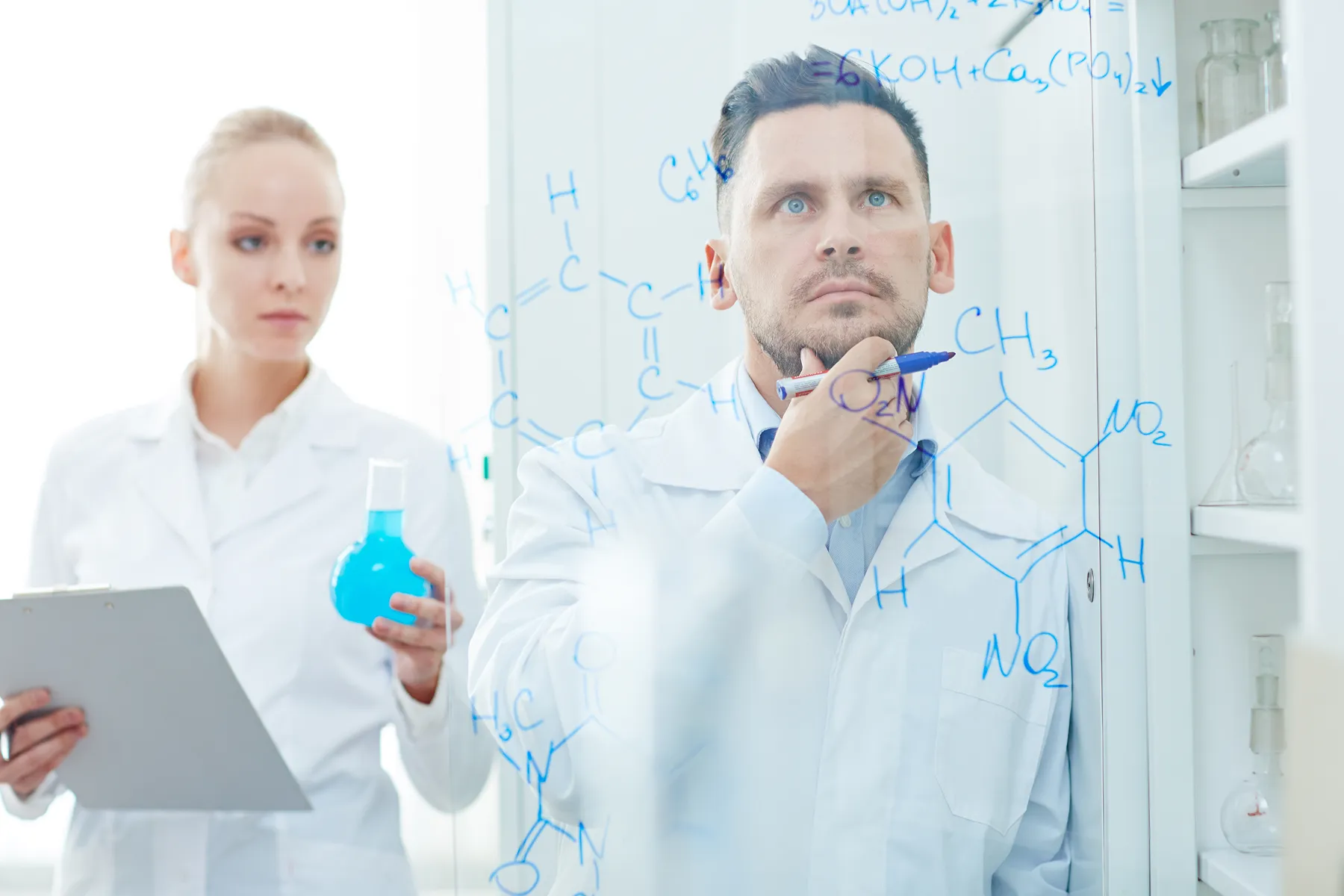 Two scientists in lab coats analyzing chemical formulas written on a transparent board, one holding a pen and the other a flask with blue liquid.