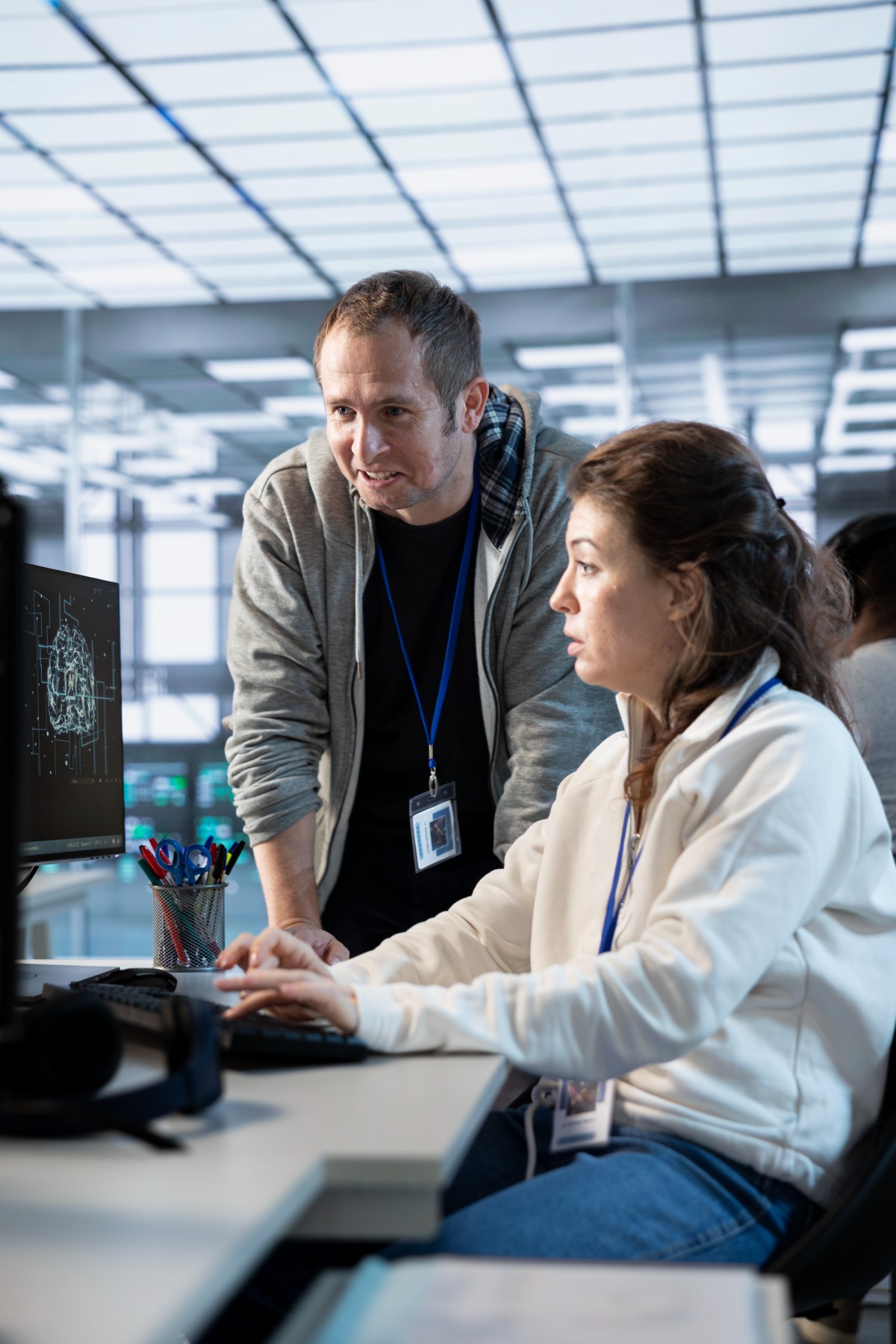 Two coworkers with ID badges collaborating at a computer workstation displaying a digital brain image.
