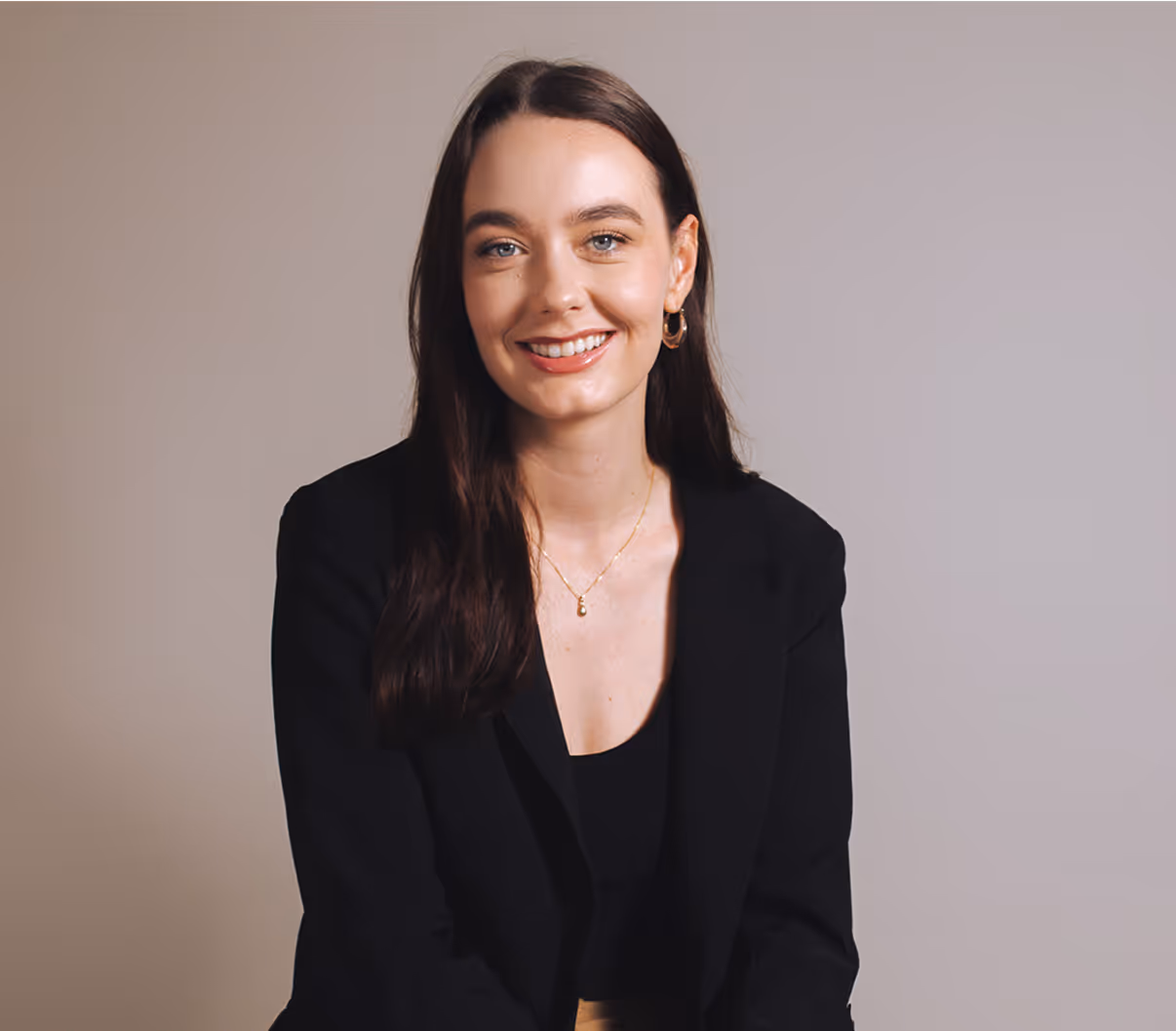 Smiling woman with long dark hair wearing a black blazer, a gold necklace, and gold hoop earrings against a plain background.