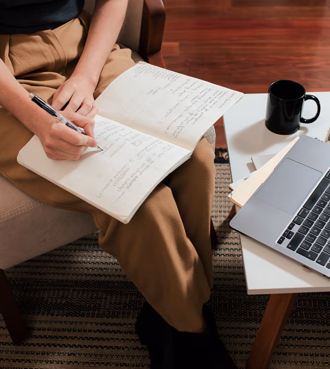 Person in brown pants writing notes in an open notebook while sitting on a chair next to a table with a laptop and black coffee mug.