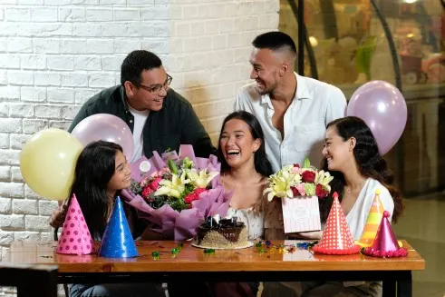A group of five friends celebrating a birthday with balloons, party hats, flowers, and a cake, smiling and enjoying together.
