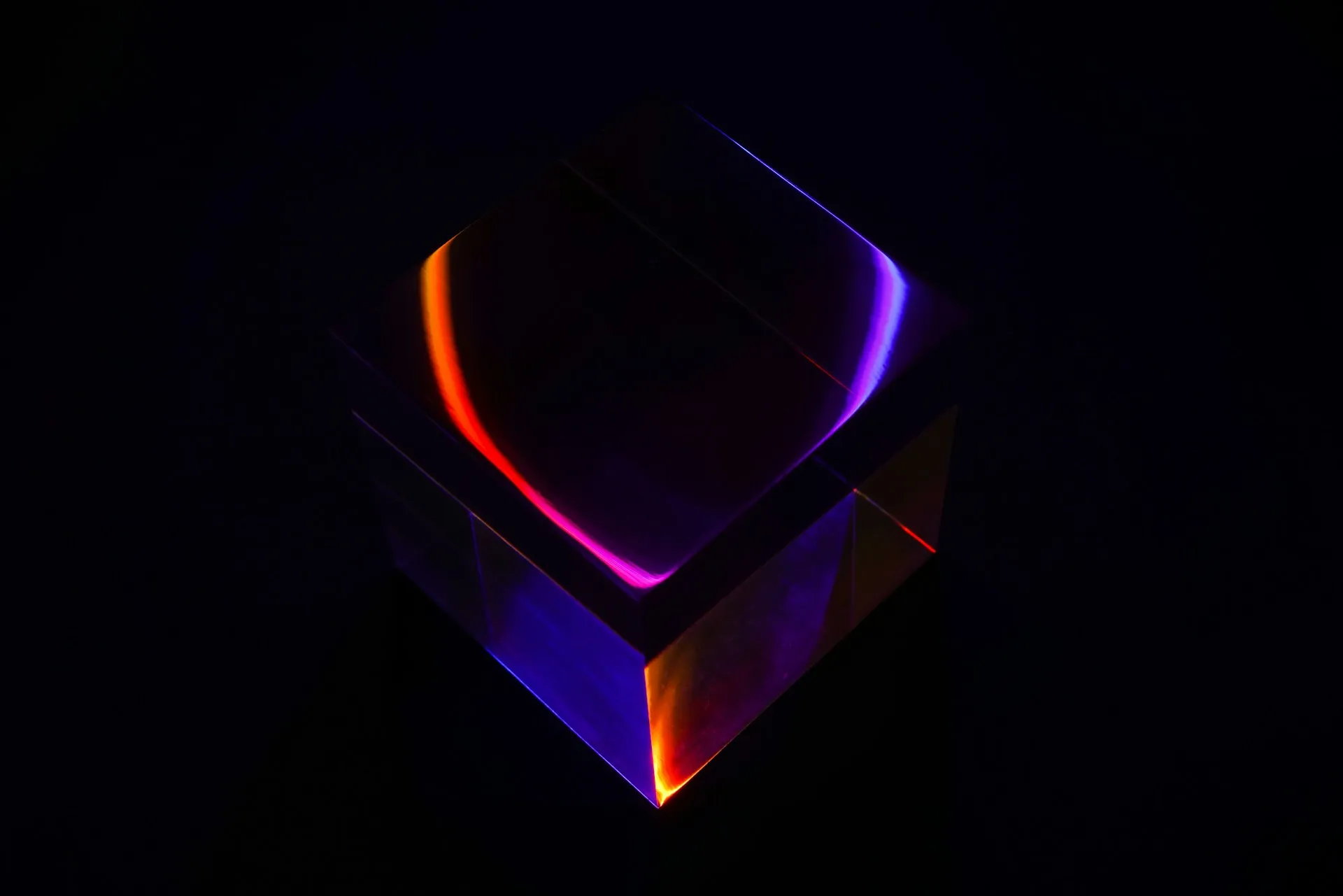 A transparent glass cube illuminated by colorful neon light reflections in shades of purple, blue, and orange against a dark background.