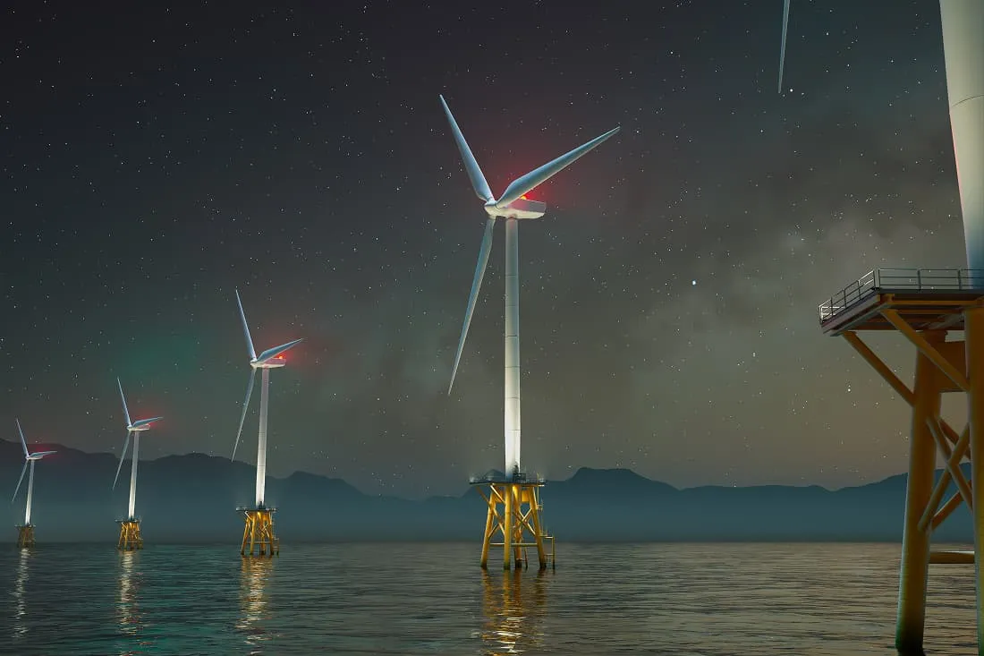 Row of offshore wind turbines with red warning lights standing in calm water under a starry night sky with mountains in the background.