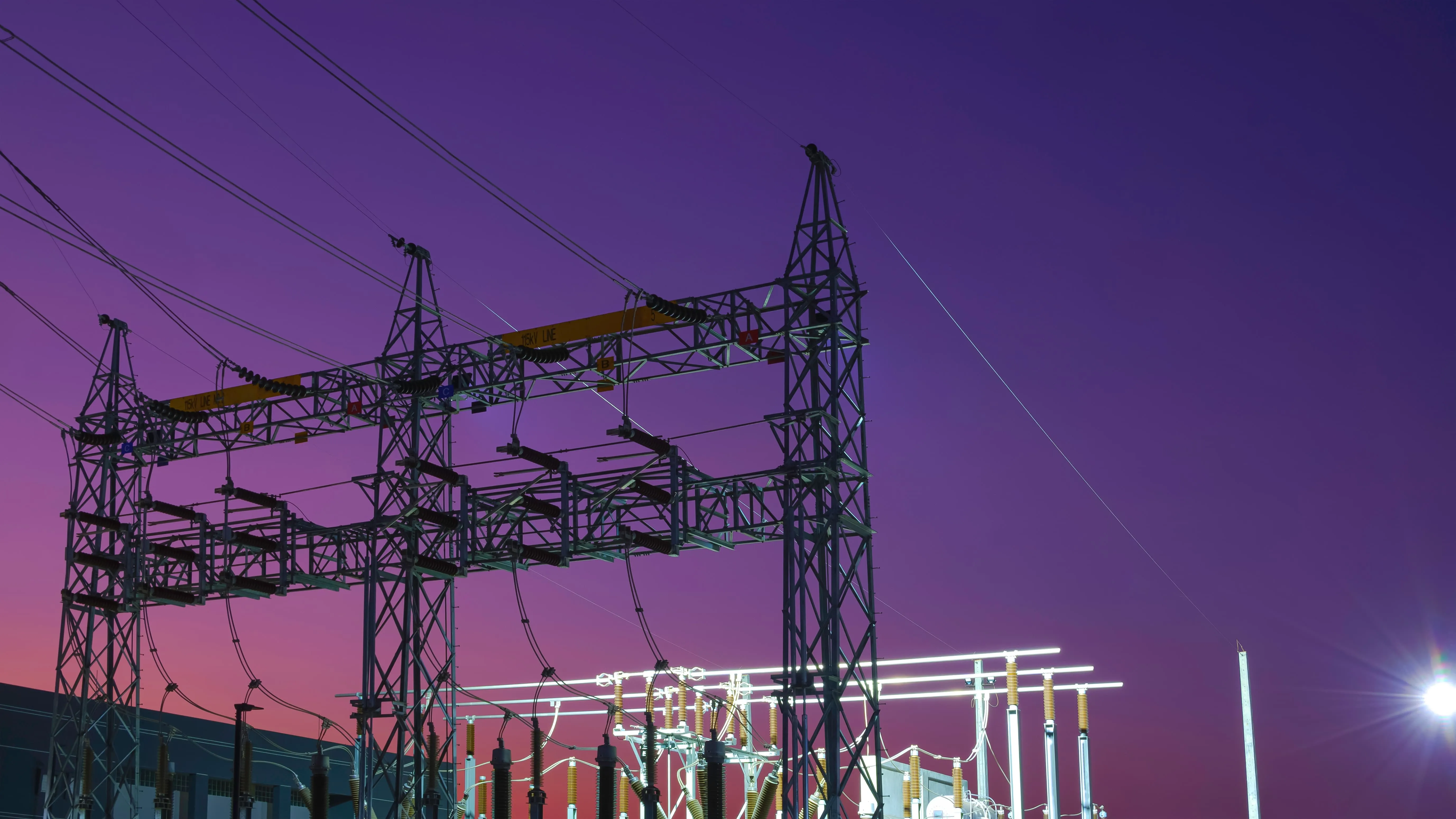 Electric power substation with tall metal frameworks and multiple cables against a purple and pink dusk sky.