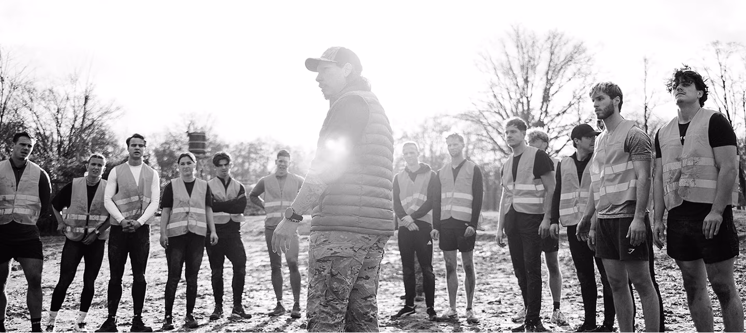 Groep jonge mensen in reflecterende hesjes luistert naar een instructeur buiten bij zonlicht.