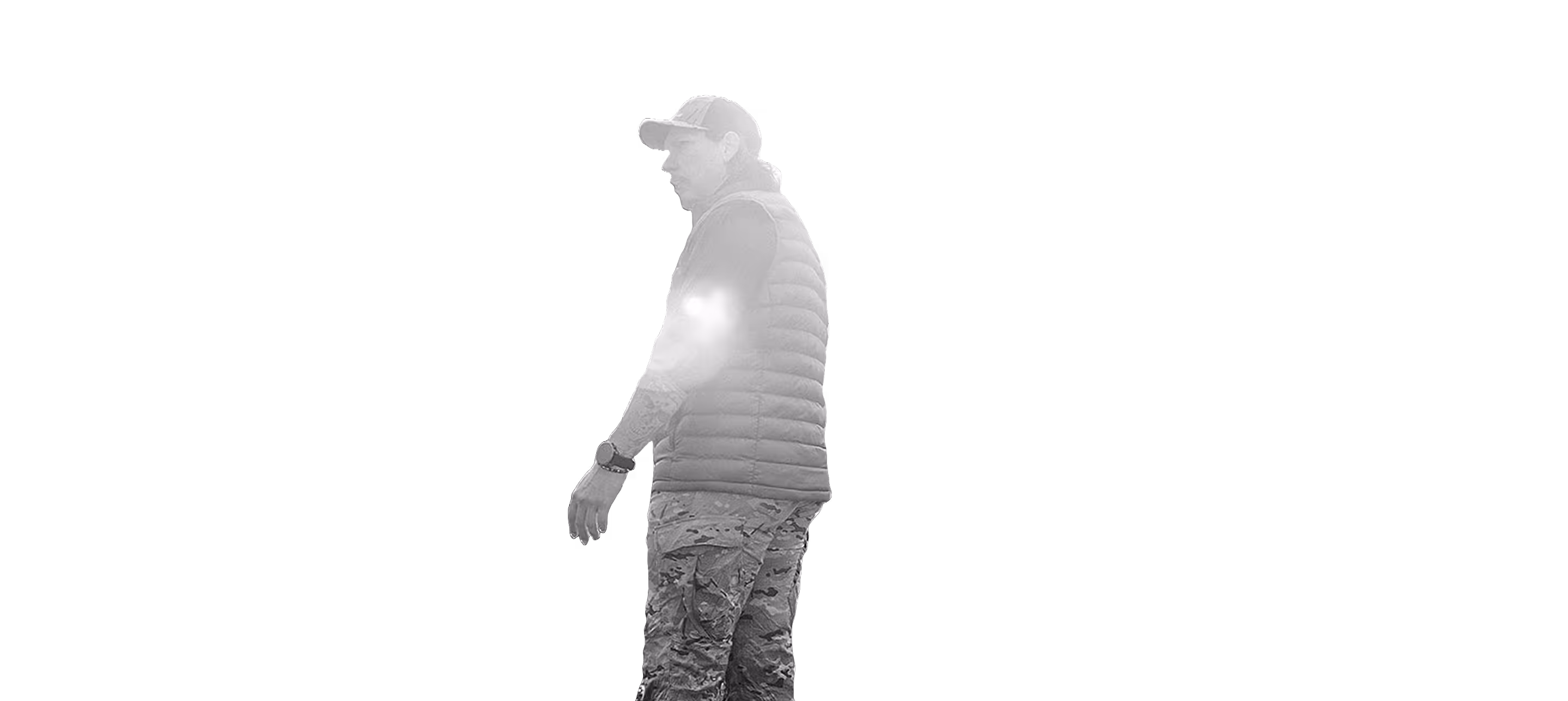 Man in camouflage pants, black puffer vest, cap, and watch, standing sideways on black background.