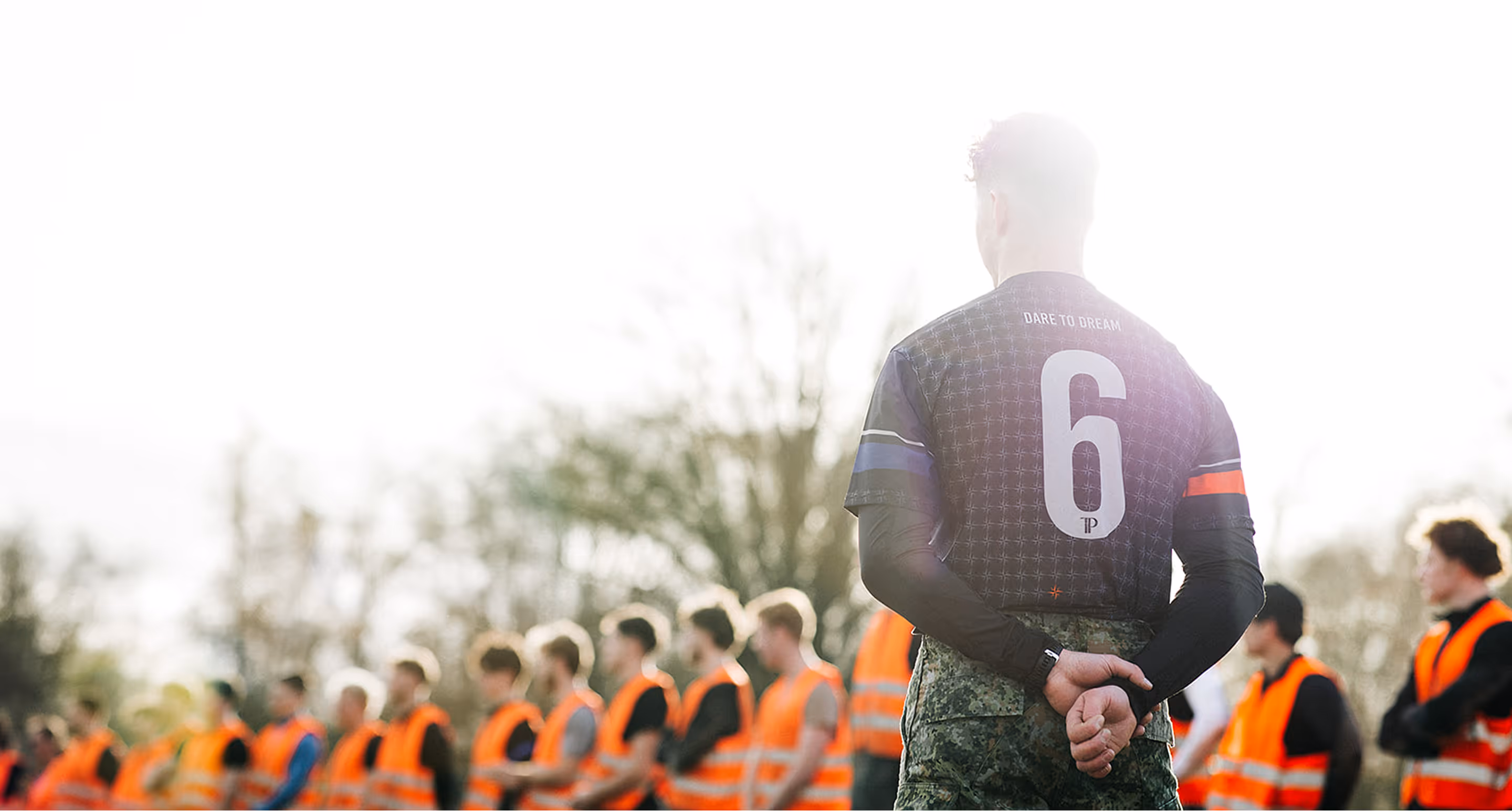 Mannen in oranje veiligheidshesjes staan in de rij; een man met 'Dare to Dream' shirt kijkt toe.