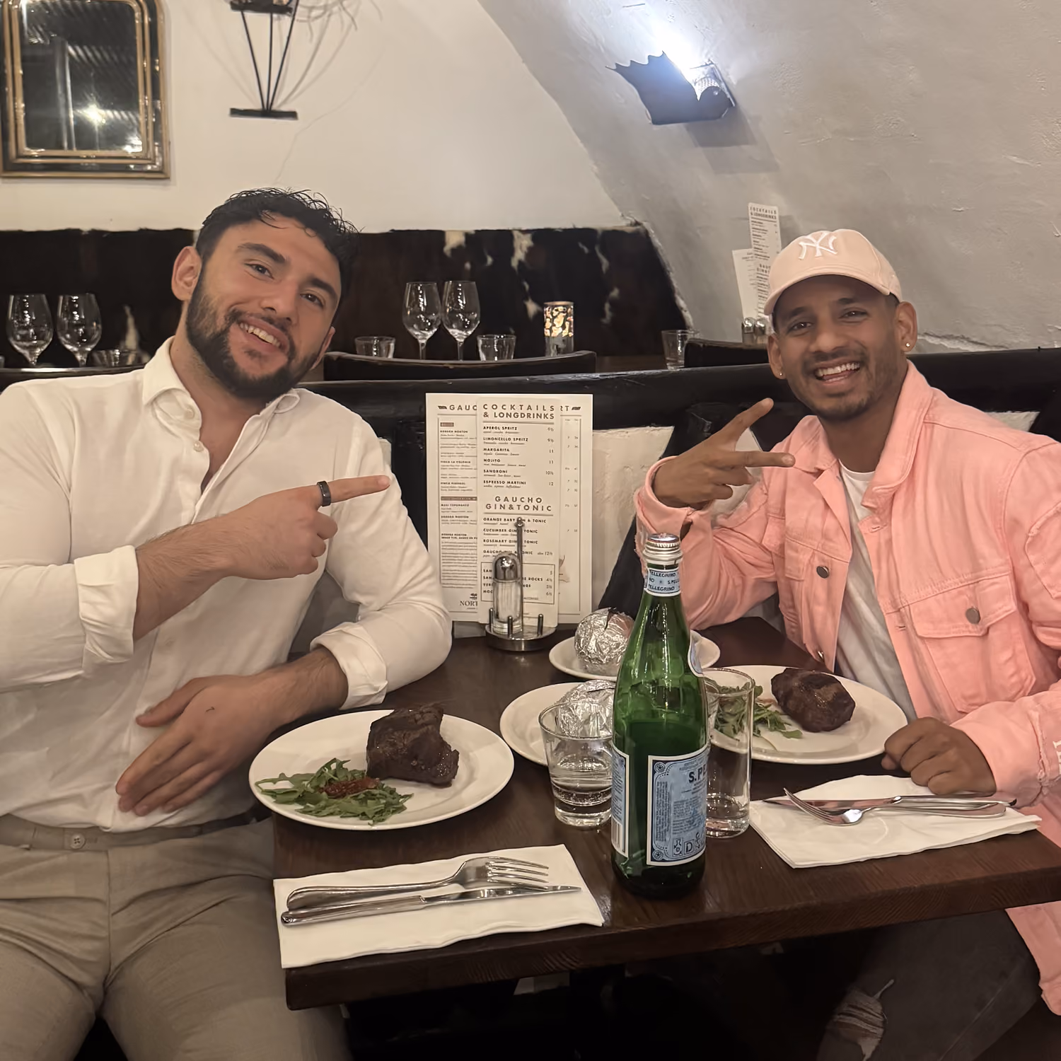 Twee mannen lachen en wijzen naar elkaar aan een tafel met biefstuk en flessen water in een restaurant.
