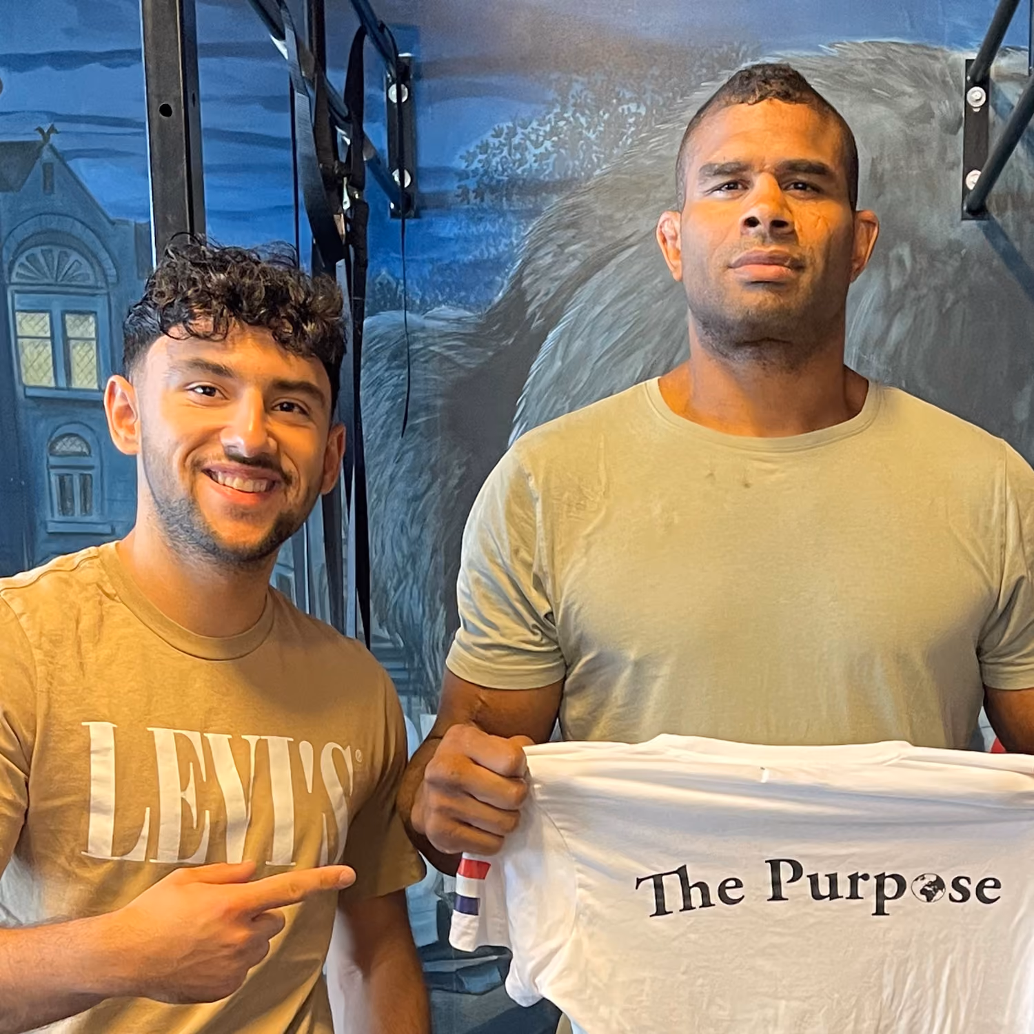 Twee mannen poseren binnen, een wijst lachend naar het shirt met 'The Purpose' tekst.