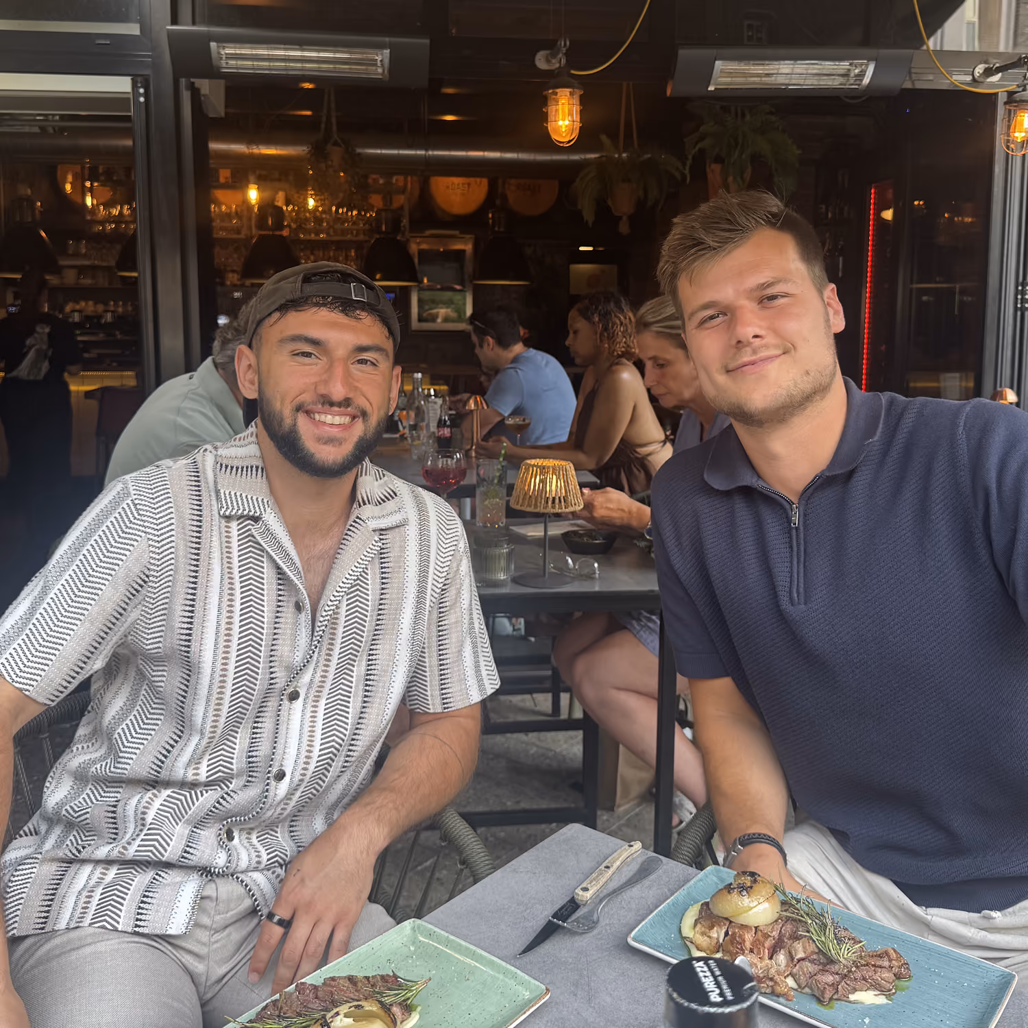 Twee mannen zitten in een restaurant samen aan tafel met borden vlees en groenten voor zich.
