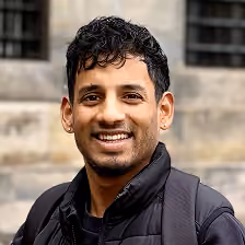 Man with short curly hair smiling outdoors wearing a black jacket and backpack.