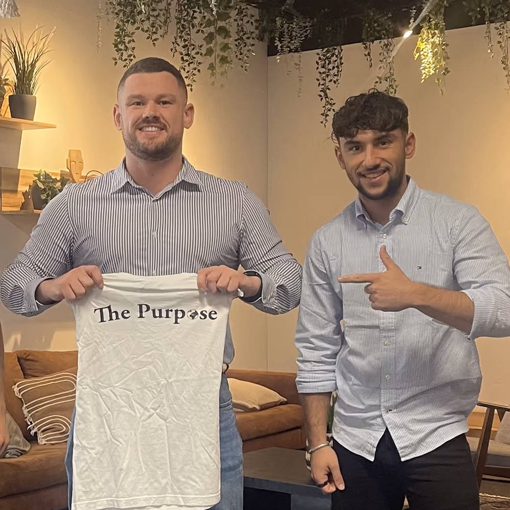 Twee mannen in overhemd, één houdt een shirt vast met tekst 'The Purpose', de ander wijst erop.