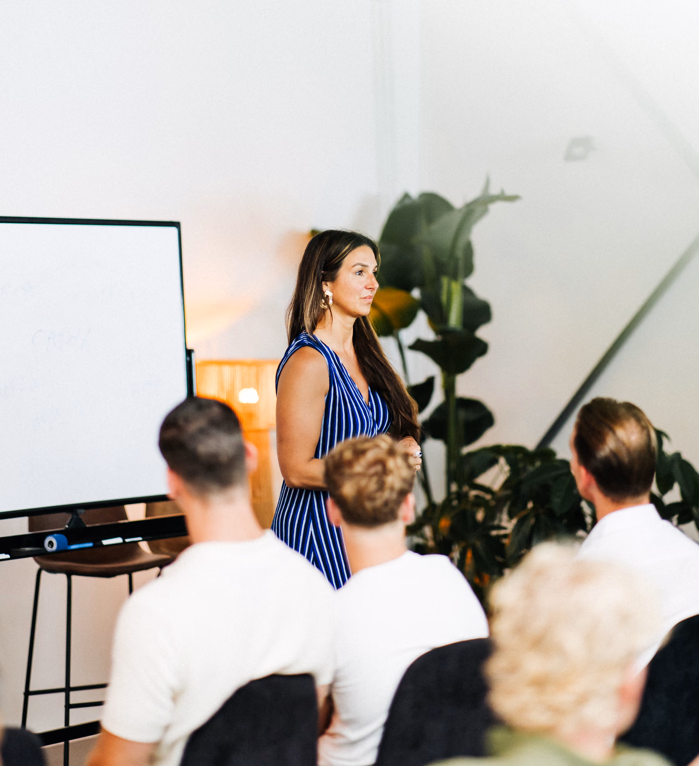 Vrouw in blauwe jurk geeft presentatie aan groep zittende mensen in lichte kamer met planten.