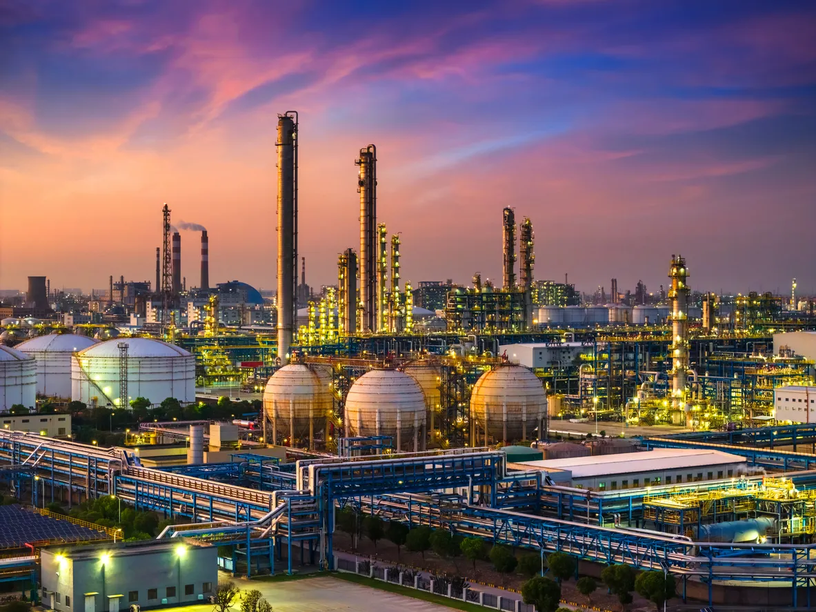 Panoramic view of a petrochemical industrial complex at sunset, featuring large storage tanks, distillation towers, and illuminated pipelines against a colorful sky.