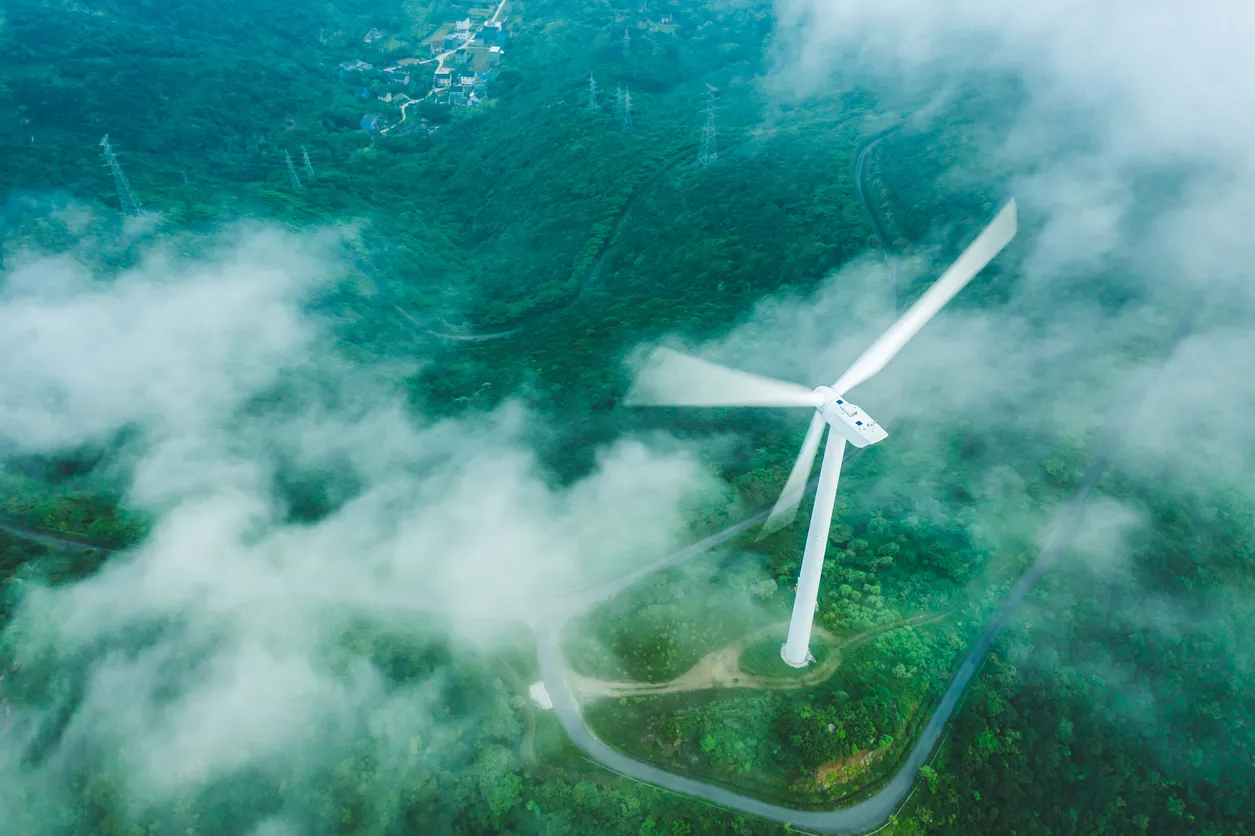 Aerial view of a wind turbine surrounded by lush green mountains and drifting fog, symbolizing clean energy generation in harmony with nature.