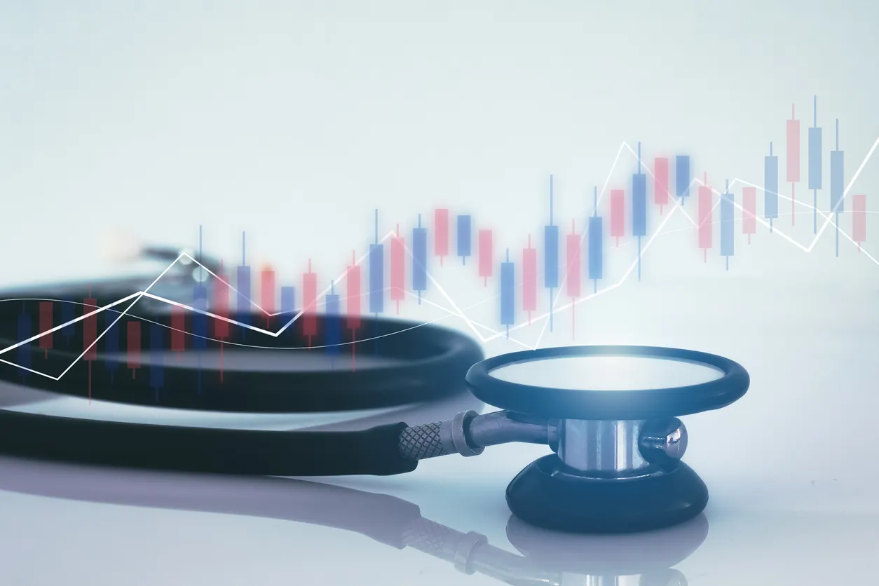 A stethoscope lies on a white surface with a superimposed financial graph in the background. The image conveys healthcare and economics interconnection.