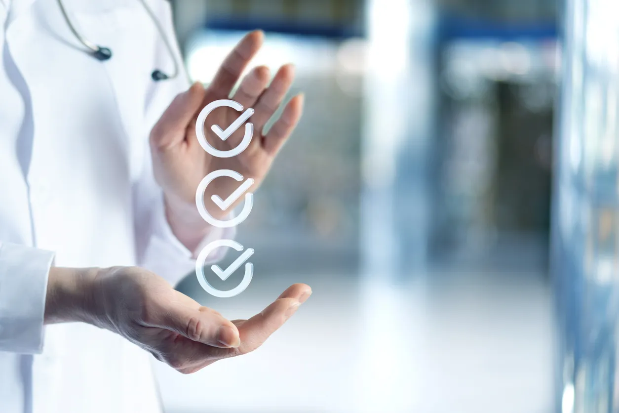 A doctor in a white coat is gesturing at three floating checkmarks, representing accomplishment and reliability, in a bright, blurred medical setting.
