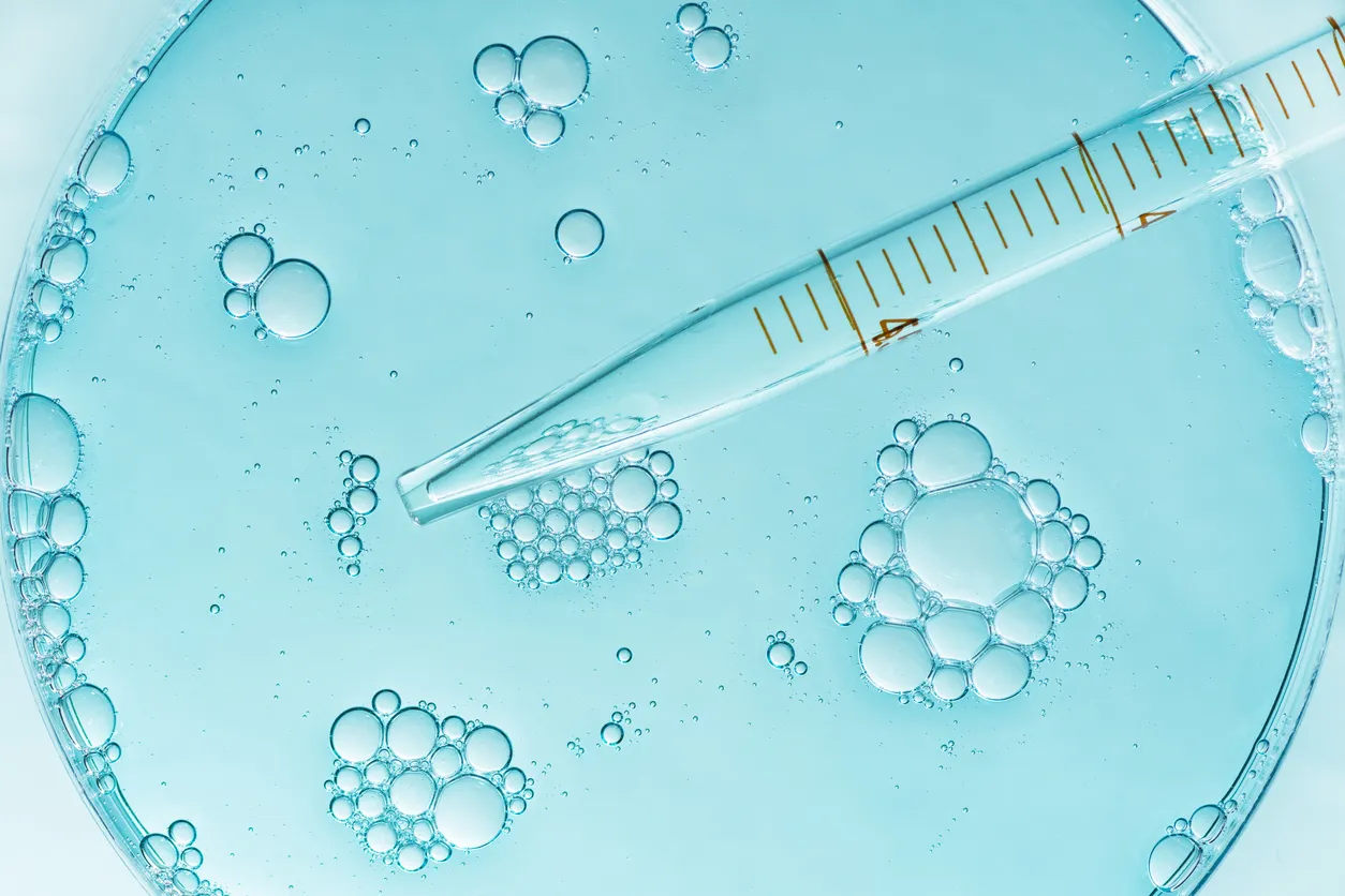 A pipette submerged in a petri dish filled with clear blue liquid and floating bubbles, illustrating a science or laboratory experiment.