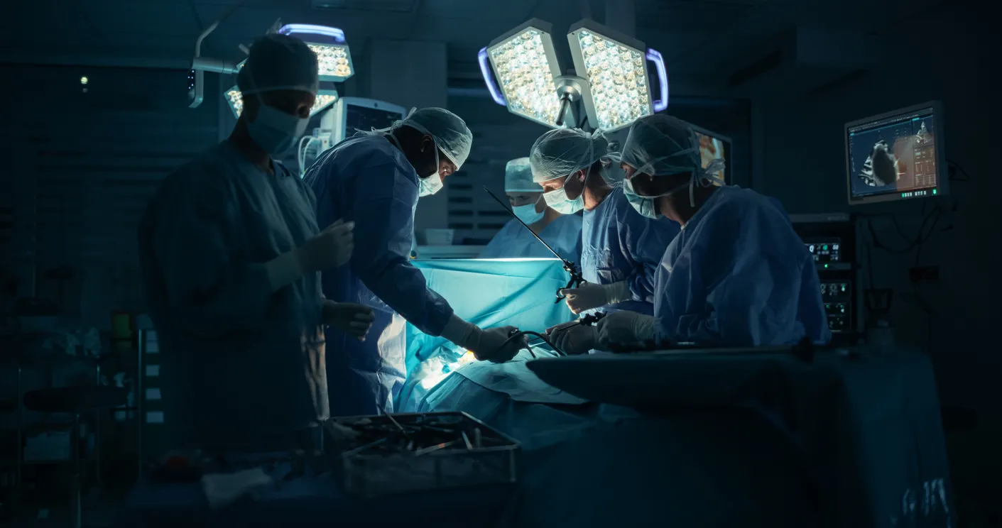 A team of surgeons in blue scrubs and masks perform surgery under bright overhead lights in an operating room. Monitors display vital signs. The mood is focused and intense.