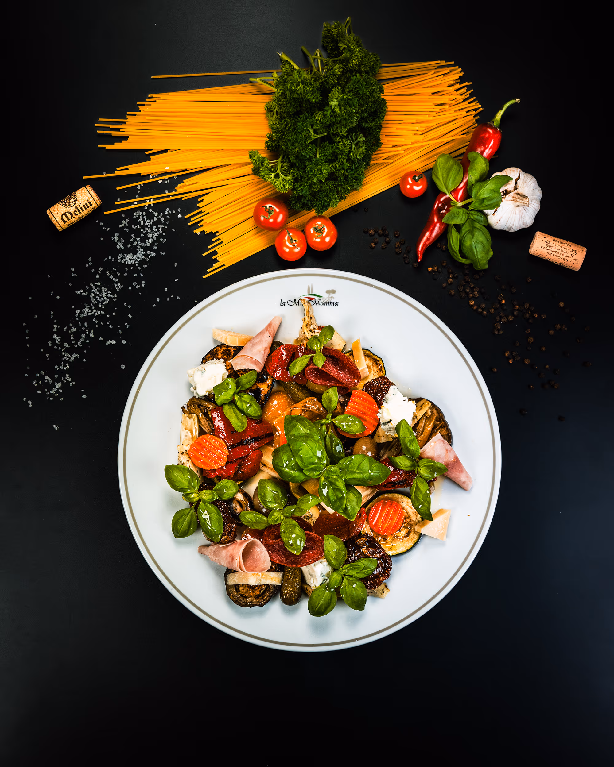 Antipasti-Teller mit Basilikum, Schinken, Salami, eingelegtem Gemüse und Käse auf weißem Teller, daneben rohe Spaghetti, Kirschtomaten, Petersilie, Knoblauch, Chilischoten, Korken und Salz auf schwarzem Hintergrund.