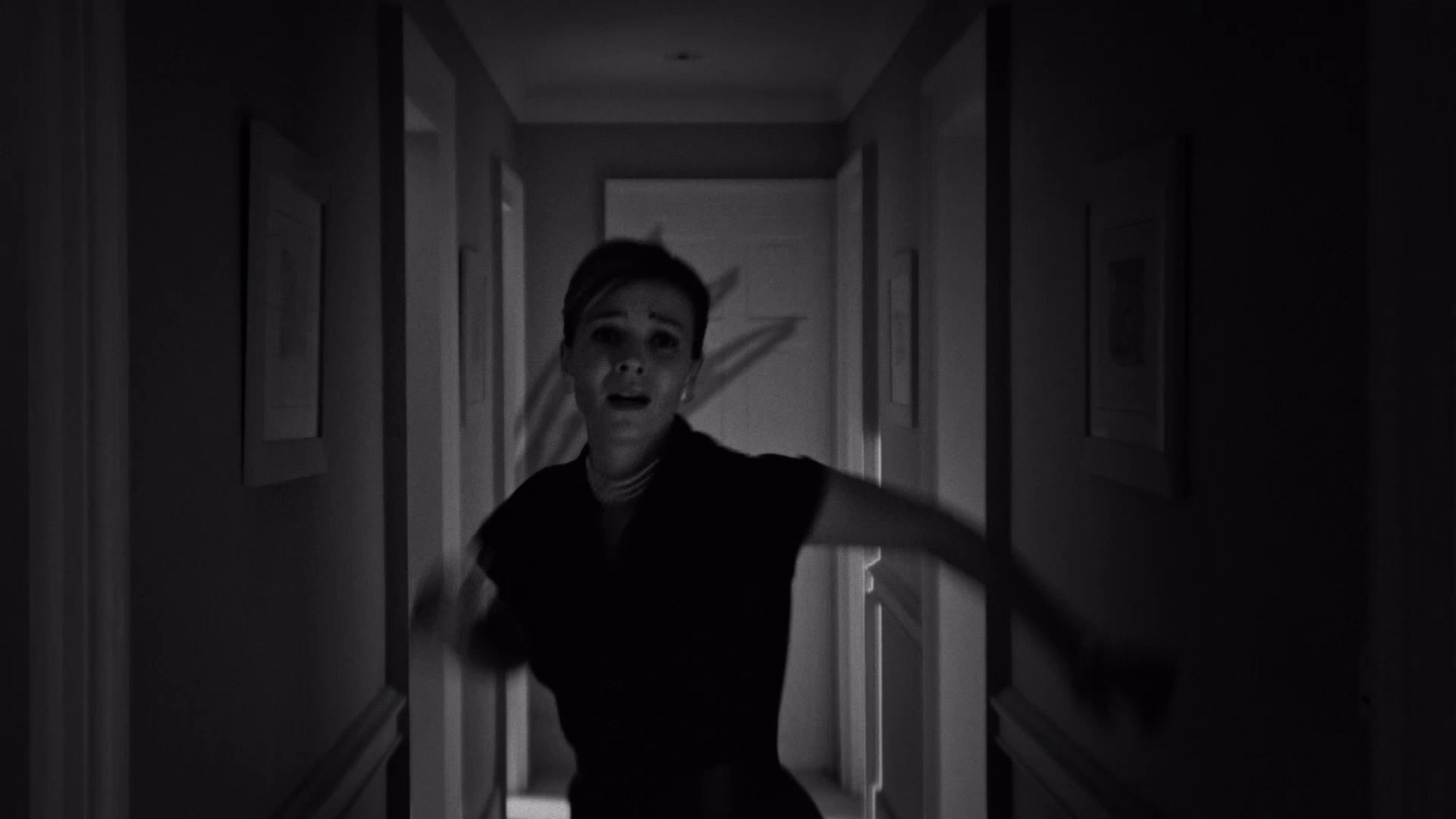 Black and white image of a woman running down a narrow hallway with a fearful expression.