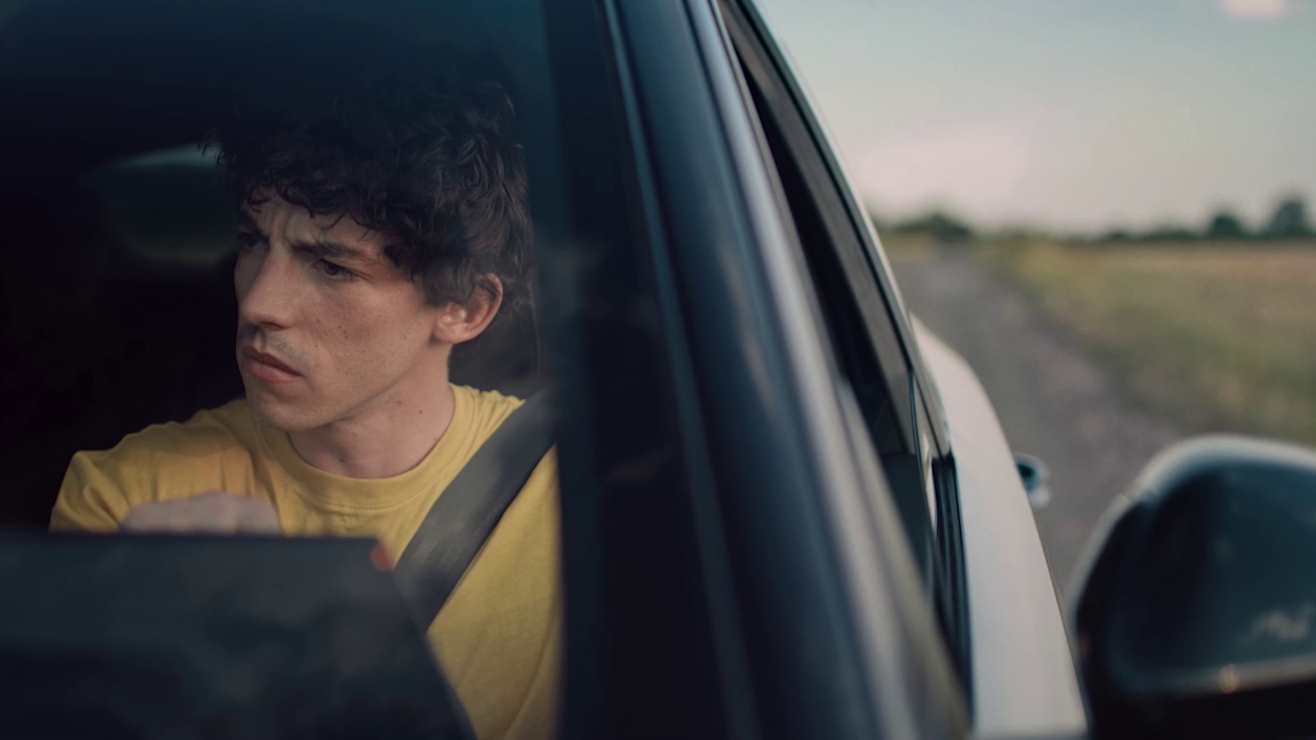 Young man in a yellow shirt driving a car and looking to the left with a serious expression.