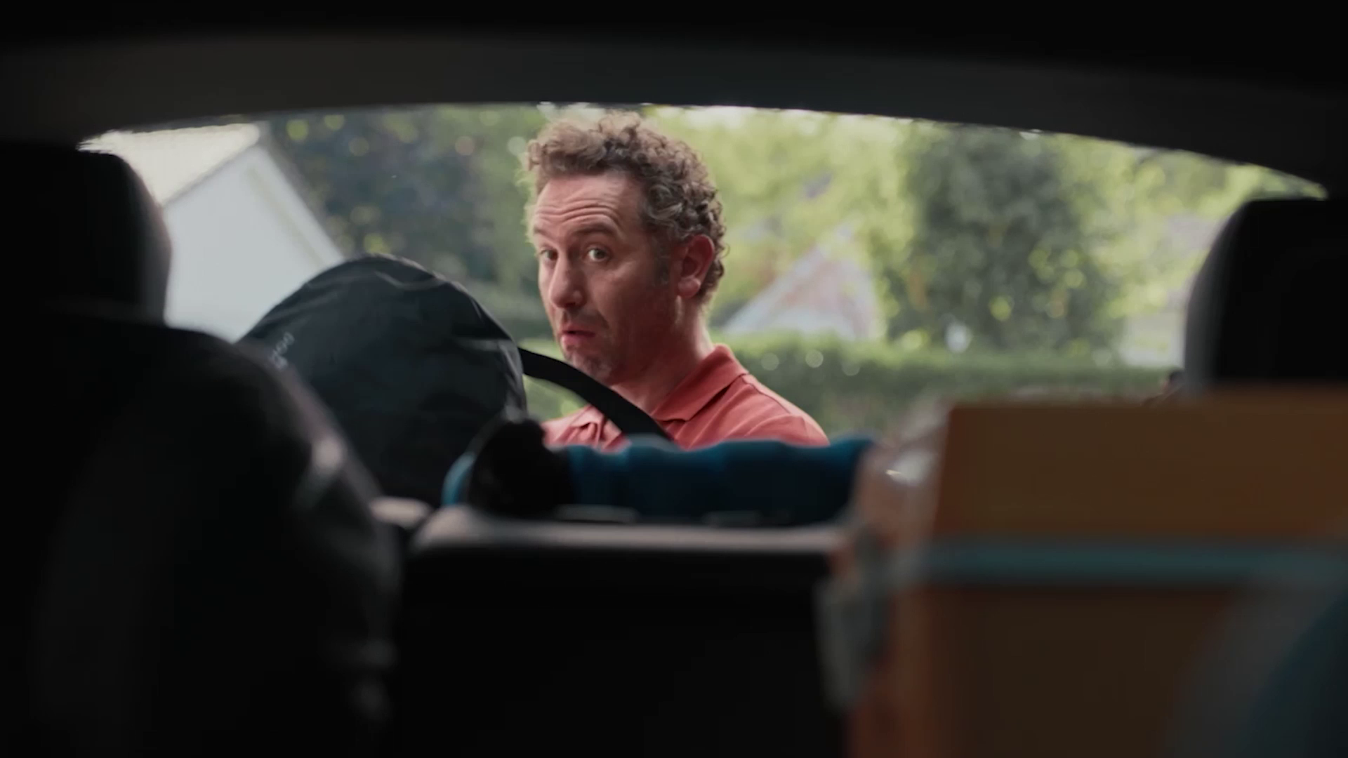 Man with curly hair looking inside a car trunk filled with bags and boxes during daytime.